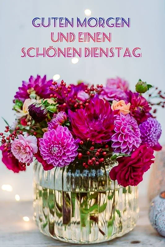Ein prachtvoller Blumenstrauß mit pinken, lila und fuchsiafarbenen Dahlien sowie roten Beeren in einer geriffelten Glasvase. Die warme Beleuchtung im Hintergrund schafft eine gemütliche und freundliche Dienstagsstimmung, perfekt für einen lieben Morgengruß.