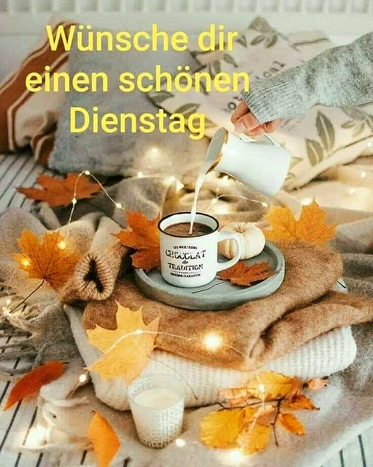 Ein herbstliches Arrangement mit einer Tasse Kaffee, die von einer Hand aus einer Kanne gefüllt wird. Die Tasse steht auf einem Teller auf einem Stapel von Pullovern. Neben dem Teller liegen Herbstblätter, eine Kerze und Lichterketten.