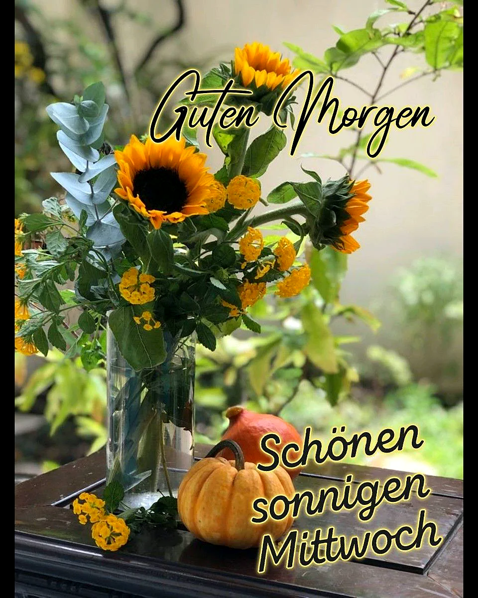 Eine Glasvase mit strahlenden Sonnenblumen, gelben Blüten und Eukalyptuszweigen steht auf dunklem Holz. Daneben liegen zwei kleine Zierkürbisse, die zusammen eine fröhliche, sonnige Morgenstimmung zur Wochenmitte vermitteln.