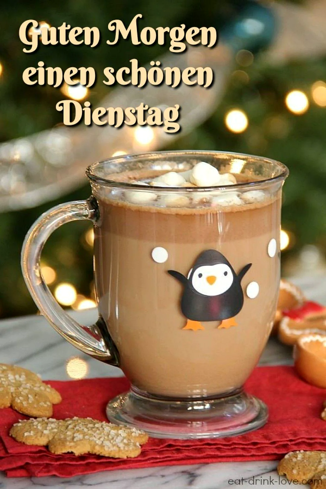 Ein Glas mit Kaffee und Marshmallows, ein süßer Pinguin ist auf dem Glas abgebildet, Kekse liegen daneben auf einem roten Tuch, im Hintergrund Weihnachtsdekoration und Lichter