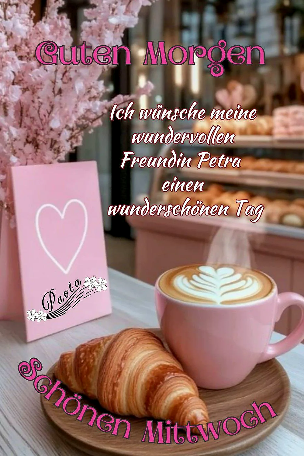 Ein rosa Kaffee-Becher mit einem Croissant auf einem Holzteller, ein rosa Kärtchen mit Herz und ein rosa Blumenstrauch im Hintergrund
