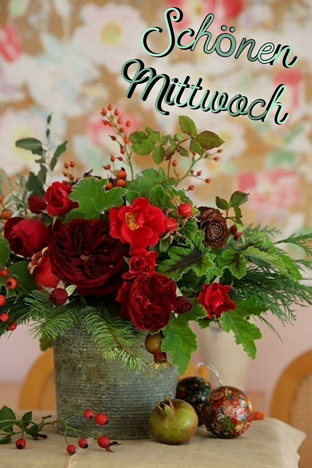 Ein Blumenstrauß mit roten Rosen und grünen Blättern in einem grauen Topf auf einem Tisch mit zwei Weihugnskugeln